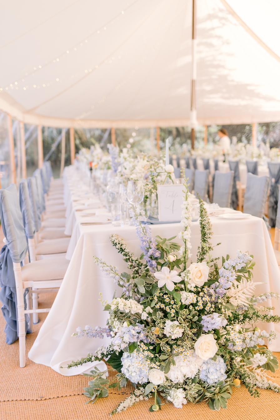 Ground arrangement of blue delphinium and white roses beside a long table, Lucia and Alex's marquee wedding in Rutland