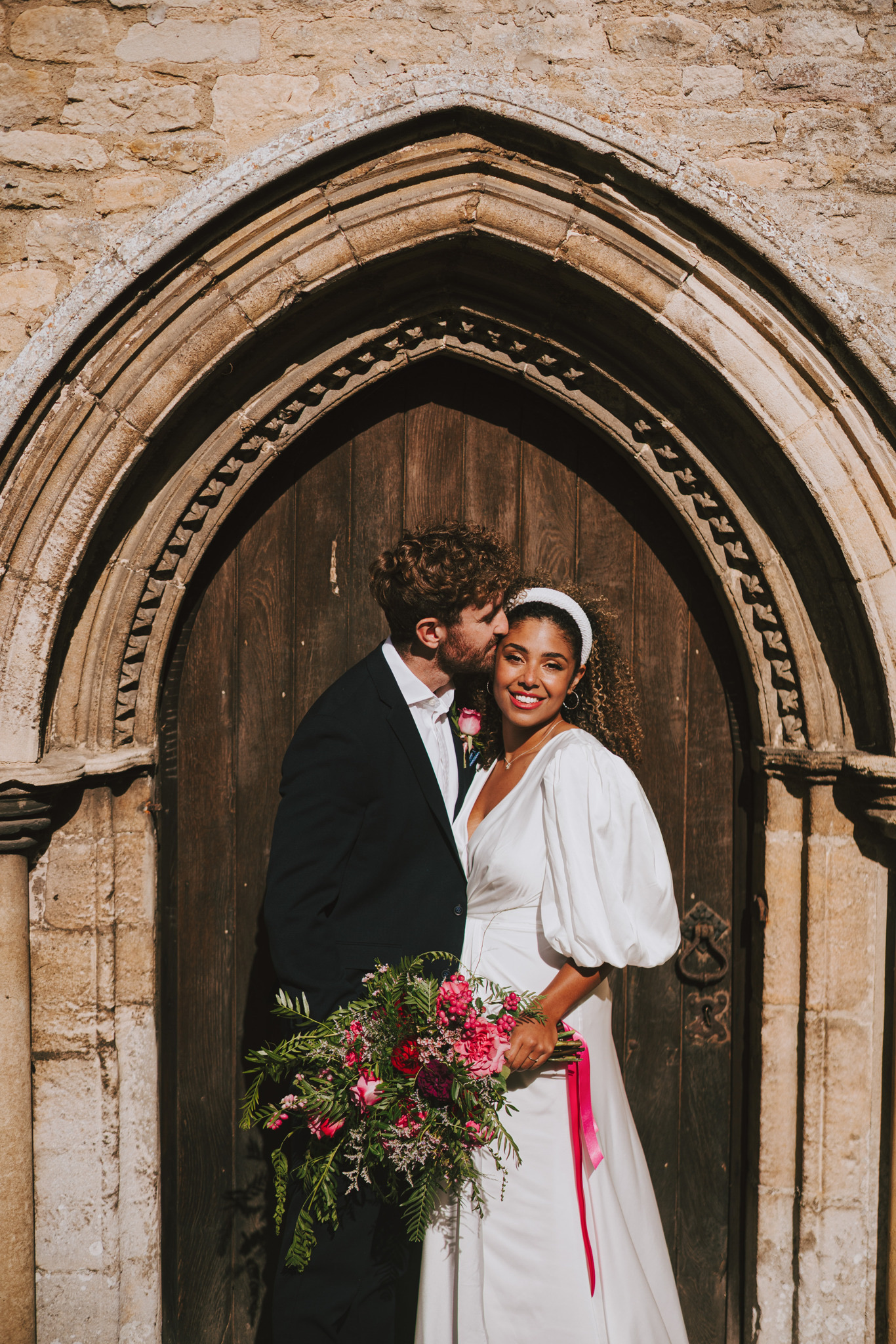 Bride and groom in the stone church doorway with a bright pink bouquet, Dionne and Alistair's manor house wedding in Lincolnshire