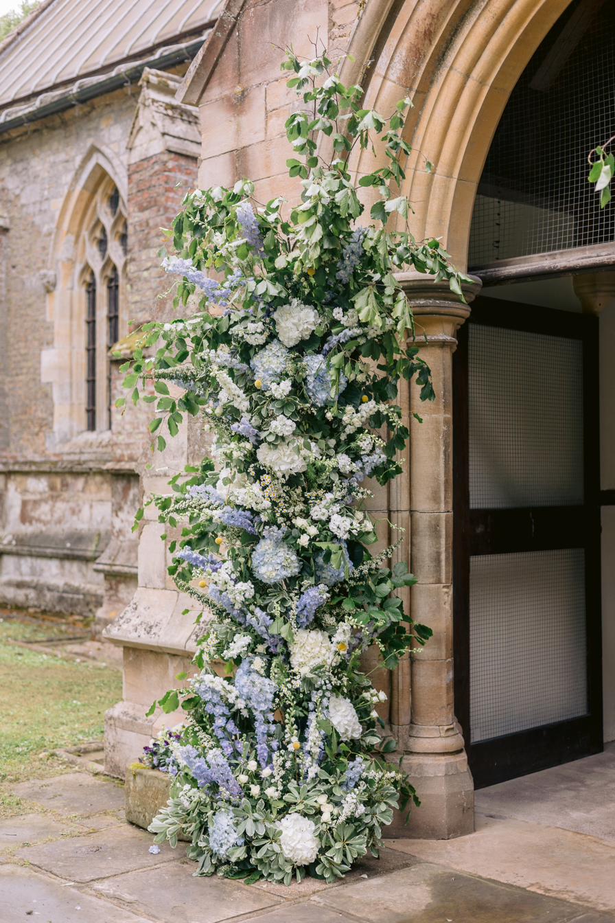 Floral column of delphinium and hydrangea at the church door, Lucia and Alex's marquee wedding in Rutland
